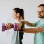 Side view of professional orthopedist in uniform and eyewear helping fit woman reaching arm with dumbbell in doctor office on blurred background